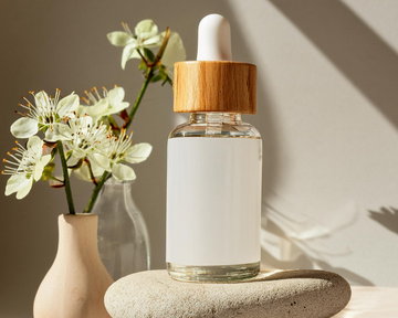 Glass dropper bottle with wooden cap on stones, next to a vase with flowers.